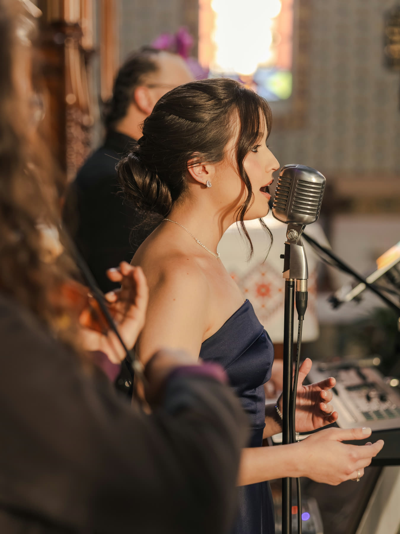 Jamilli de perfil cantando ao microfone vintage, com a luz da igreja ao fundo